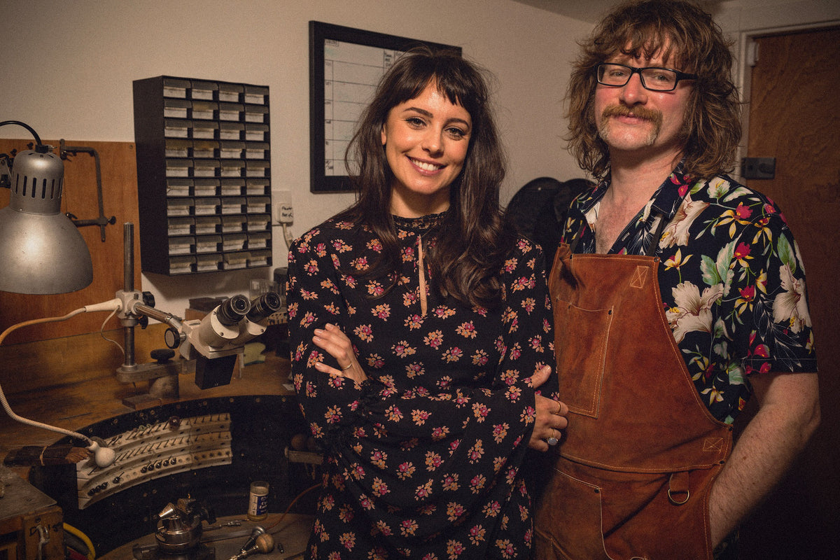 A man and a woman standing together in a workshop. The woman wears a black long-sleeve top with a pink and white floral pattern, and the man wears a colorful floral shirt and a brown leather apron.